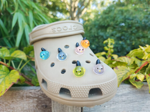 Taupe-colored Croc shoe adorned with glow-in-the-dark bird charms. Each charm features a small, round bird with a beak and black wings, glowing softly in various colors. The shoe is positioned outdoors against a backdrop of green foliage.