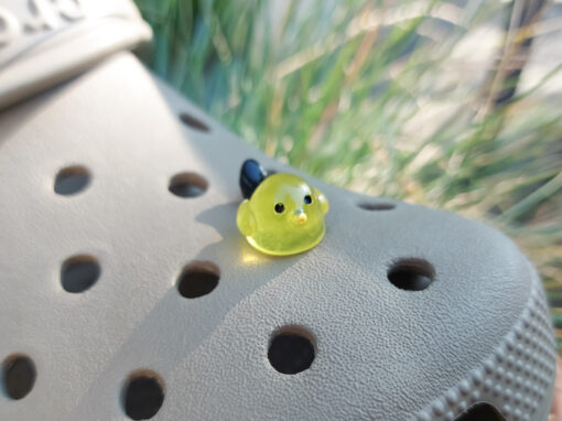 Taupe-colored Croc shoe adorned with glow-in-the-dark bird charms. Each charm features a small, round bird with a beak and black wings, glowing softly in various colors. The shoe is positioned outdoors against a backdrop of green foliage.