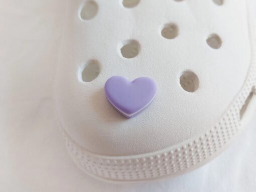 Yellow Crocs adorned with pastel-colored macaron heart charms, placed among vibrant pink flowers. The charms add a playful and loving touch to the shoes.