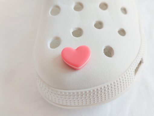 Yellow Crocs adorned with pastel-colored macaron heart charms, placed among vibrant pink flowers. The charms add a playful and loving touch to the shoes.