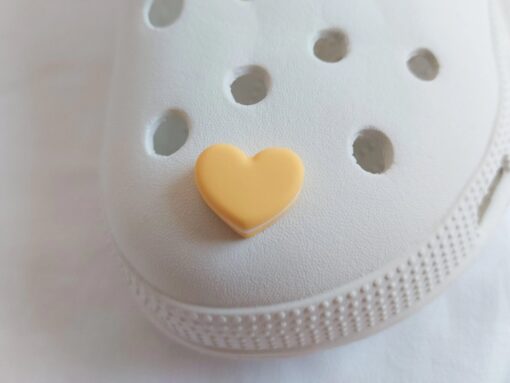 Yellow Crocs adorned with pastel-colored macaron heart charms, placed among vibrant pink flowers. The charms add a playful and loving touch to the shoes.