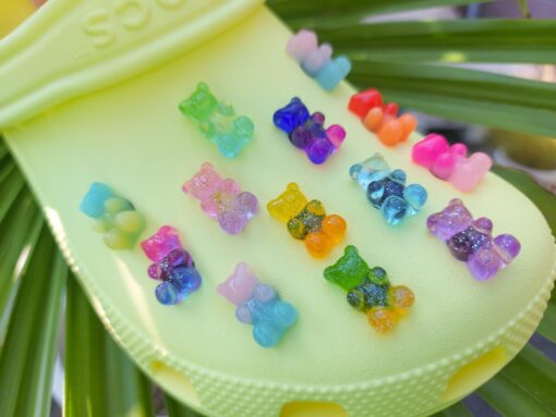 Bright yellow Croc shoe decorated with colorful gummy bear-shaped charms, each in various shades of blue, pink, orange, purple, green, and more. The gummy bear charms sparkle under the sunlight, sitting on a green leaf background.