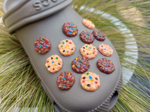 Kaki Crocs decorated with various cookie charms, featuring chocolate chip and candy-topped cookies, displayed on a backdrop of green grass.