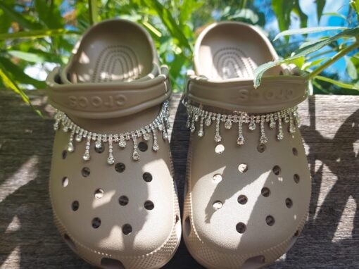 Close-up of a rhinestone chain charm draped across a pair of Crocs, featuring sparkling rhinestones that catch the light. The chain is securely attached, adding a chic and luxurious touch to the shoes with its elegant, shimmering design.