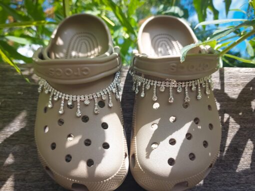 Close-up of a rhinestone chain charm draped across a pair of Crocs, featuring sparkling rhinestones that catch the light. The chain is securely attached, adding a chic and luxurious touch to the shoes with its elegant, shimmering design.