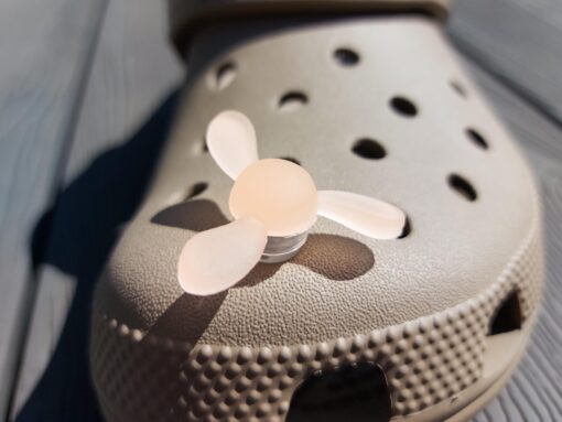 Close-up of glow-in-the-dark windmill croc charms attached to a beige Croc, displayed outdoors in sunlight. The colorful windmill blades spin as you walk, adding a fun, dynamic feature to your shoes.