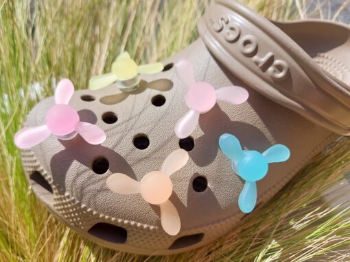 Close-up of glow-in-the-dark windmill croc charms attached to a beige Croc, displayed outdoors in sunlight. The colorful windmill blades spin as you walk, adding a fun, dynamic feature to your shoes.