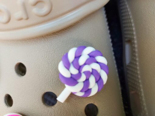 Pair of beige Crocs adorned with colorful lollipop charms, each charm featuring a different candy swirl pattern. The Crocs are displayed on a green palm leaf, with the bright lollipops adding a fun, sweet, and whimsical touch to the footwear.