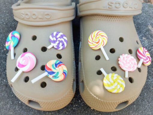 Pair of beige Crocs adorned with colorful lollipop charms, each charm featuring a different candy swirl pattern. The Crocs are displayed on a green palm leaf, with the bright lollipops adding a fun, sweet, and whimsical touch to the footwear.
