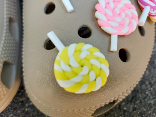 Pair of beige Crocs adorned with colorful lollipop charms, each charm featuring a different candy swirl pattern. The Crocs are displayed on a green palm leaf, with the bright lollipops adding a fun, sweet, and whimsical touch to the footwear.