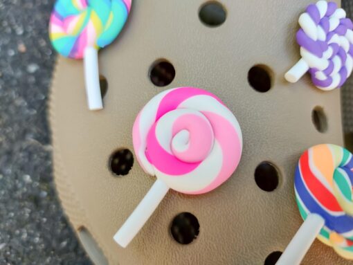 Pair of beige Crocs adorned with colorful lollipop charms, each charm featuring a different candy swirl pattern. The Crocs are displayed on a green palm leaf, with the bright lollipops adding a fun, sweet, and whimsical touch to the footwear.