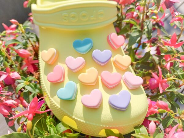 Yellow Crocs adorned with pastel-colored macaron heart charms, placed among vibrant pink flowers. The charms add a playful and loving touch to the shoes.