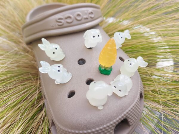 A set of 7 unique glow-in-the-dark bunny charms and 1 carrot charm arranged on a shoe. The bunnies are in various poses, and the carrot sits in the center, all glowing softly in a darkened setting.