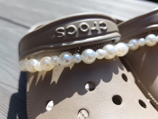 Close-up of a white pearl chain charm on a pair of Crocs, featuring a string of lustrous pearls elegantly draped across the shoe. The charm adds a classic and timeless touch to the footwear, creating a sophisticated and luxurious look.