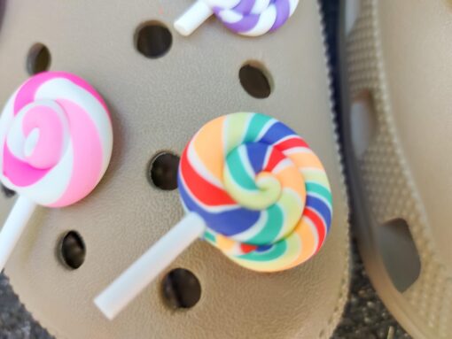 Pair of beige Crocs adorned with colorful lollipop charms, each charm featuring a different candy swirl pattern. The Crocs are displayed on a green palm leaf, with the bright lollipops adding a fun, sweet, and whimsical touch to the footwear.