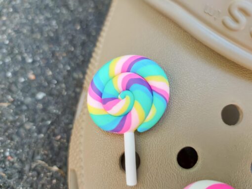 Pair of beige Crocs adorned with colorful lollipop charms, each charm featuring a different candy swirl pattern. The Crocs are displayed on a green palm leaf, with the bright lollipops adding a fun, sweet, and whimsical touch to the footwear.
