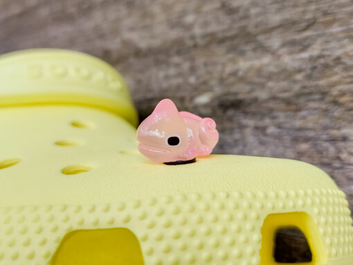 A close-up of a yellow Croc shoe with vibrant glow-in-the-dark chameleon charms in different colors (green, orange, pink, blue, and yellow) displayed outdoors on green grass.