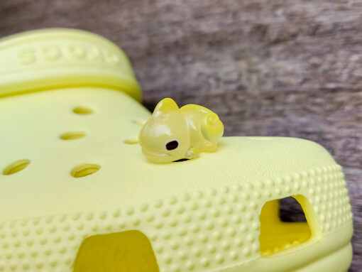 A close-up of a yellow Croc shoe with vibrant glow-in-the-dark chameleon charms in different colors (green, orange, pink, blue, and yellow) displayed outdoors on green grass.
