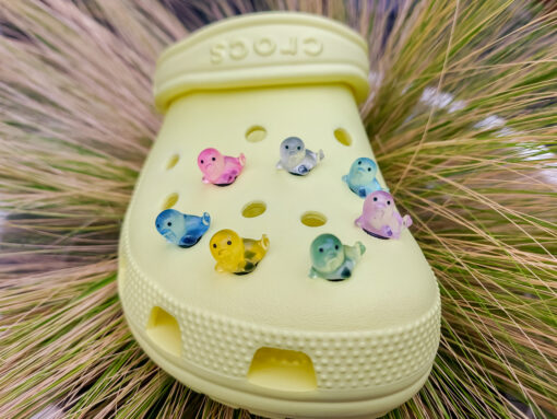 A yellow Croc shoe adorned with colorful glow-in-the-dark seal charms in shades of pink, blue, green, yellow, and purple, set against a natural background with blades of grass.
