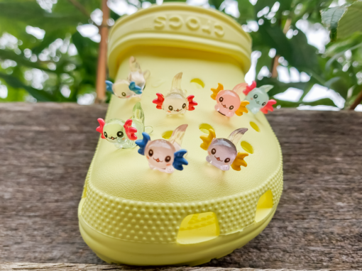 A yellow Croc shoe featuring colorful glow-in-the-dark axolotl charms in shades of green, pink, blue, orange, and silver, displayed outdoors on a wooden surface. The charms, with smiling faces and frilly gills, brighten the shoe.
