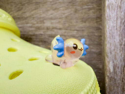 A yellow Croc shoe featuring colorful glow-in-the-dark axolotl charms in shades of green, pink, blue, orange, and silver, displayed outdoors on a wooden surface. The charms, with smiling faces and frilly gills, brighten the shoe.