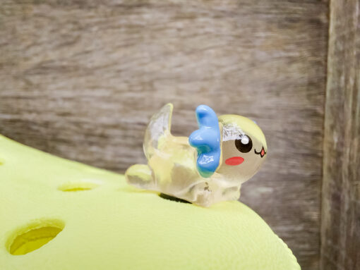 A yellow Croc shoe featuring colorful glow-in-the-dark axolotl charms in shades of green, pink, blue, orange, and silver, displayed outdoors on a wooden surface. The charms, with smiling faces and frilly gills, brighten the shoe.