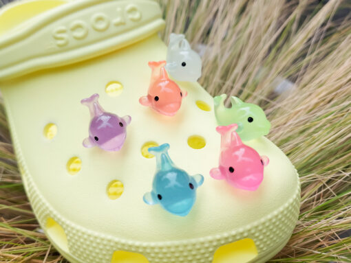 Six glow-in-the-dark dolphin charms in various pastel colors, including purple, pink, orange, green, blue, and white, are arranged on a yellow Crocs shoe. The dolphins have tiny black eyes and wide grins, giving them a playful and cute appearance.