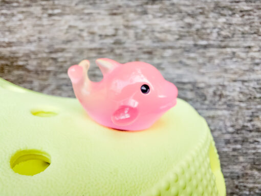 Six glow-in-the-dark dolphin charms in various pastel colors, including purple, pink, orange, green, blue, and white, are arranged on a yellow Crocs shoe. The dolphins have tiny black eyes and wide grins, giving them a playful and cute appearance.