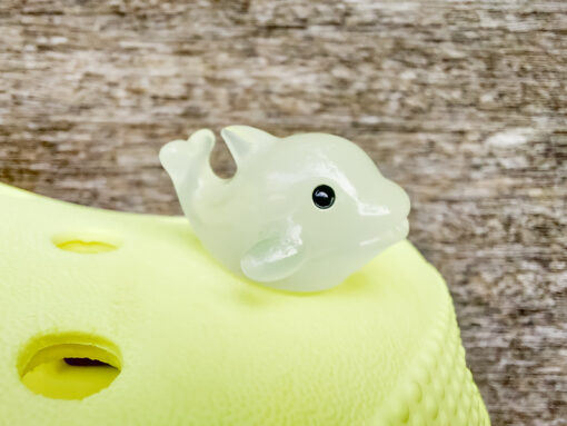 Six glow-in-the-dark dolphin charms in various pastel colors, including purple, pink, orange, green, blue, and white, are arranged on a yellow Crocs shoe. The dolphins have tiny black eyes and wide grins, giving them a playful and cute appearance.