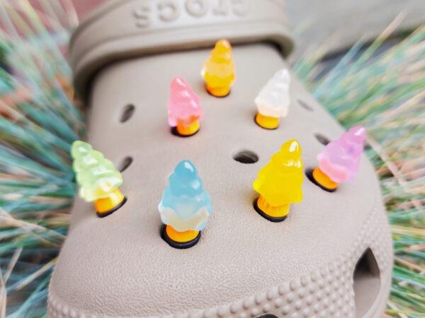 A beige Crocs shoe adorned with small, glowing Christmas tree charms in a variety of soft colors, including yellow, pink, green, and blue. Each tree charm has a luminous effect, creating a festive and playful look.