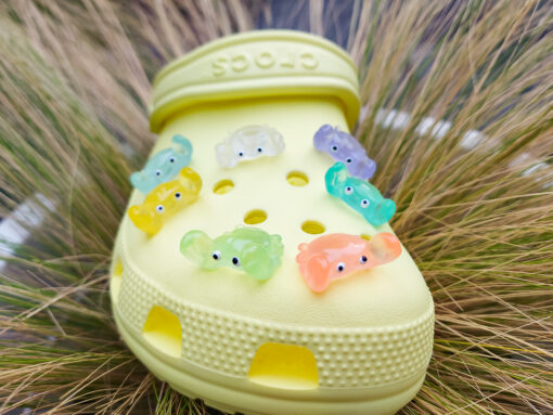 Close-up of a yellow Croc shoe adorned with colorful glow-in-the-dark crab charms in various shades. The charms have cute, detailed faces and are displayed in vibrant colors, ready to glow once charged under sunlight.