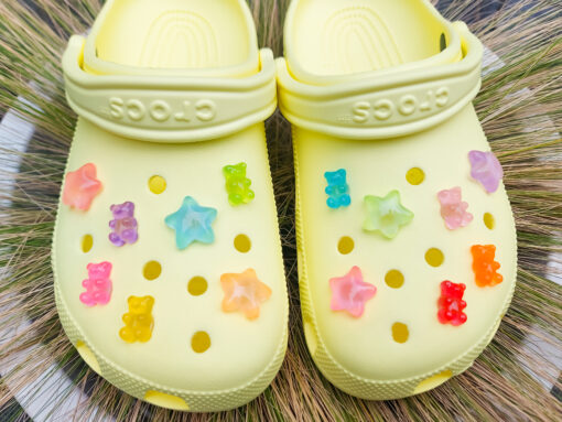 Close-up of yellow Crocs featuring 14 colorful glow-in-the-dark gummy bear and star charms in shades of red, orange, pink, green, purple, and blue, arranged on the shoes, glowing softly in daylight against the bright background.