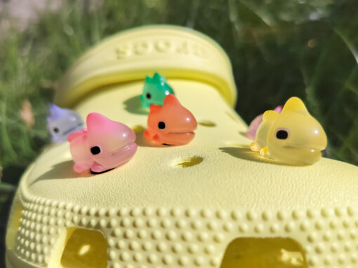 A close-up of a yellow Croc shoe with vibrant glow-in-the-dark chameleon charms in different colors (green, orange, pink, blue, and yellow) displayed outdoors on green grass.