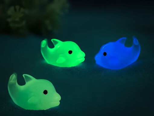 Six glow-in-the-dark dolphin charms in various pastel colors, including purple, pink, orange, green, blue, and white, are arranged on a yellow Crocs shoe. The dolphins have tiny black eyes and wide grins, giving them a playful and cute appearance.