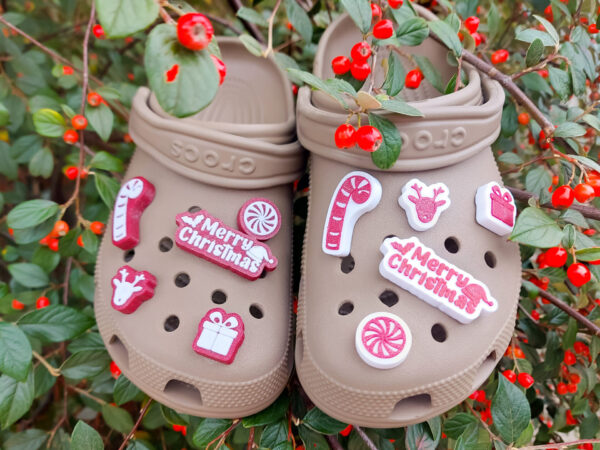 Tan Crocs adorned with festive 3D printed Christmas-themed charms, including candy canes, reindeer, gifts, and Merry Christmas text, surrounded by bright red berries and green leaves.