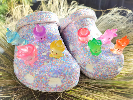 A pair of glittery Crocs adorned with colorful candle-shaped charms in shades of pink, green, blue, and yellow, set against a soft natural background.