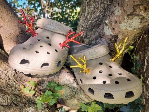 A pair of tan Crocs decorated with red and gold reindeer antler charms, perched on a tree branch, perfect for holiday-themed footwear decorations. Available in multiple festive colors.