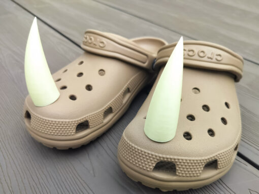 Pair of tan Crocs with large glow-in-the-dark 3D printed rhino horn charms attached to the front, placed on a wooden surface.