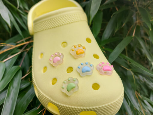 Glow-in-the-dark pastel paw print charms in pink, yellow, blue, and green, attached to a yellow shoe. The charms have a soft glow effect and add a playful animal-inspired look.