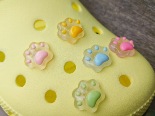 Glow-in-the-dark pastel paw print charms in pink, yellow, blue, and green, attached to a yellow shoe. The charms have a soft glow effect and add a playful animal-inspired look.