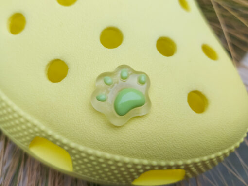 Glow-in-the-dark pastel paw print charms in pink, yellow, blue, and green, attached to a yellow shoe. The charms have a soft glow effect and add a playful animal-inspired look.