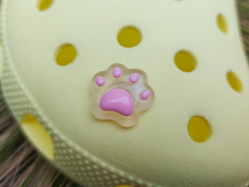 Glow-in-the-dark pastel paw print charms in pink, yellow, blue, and green, attached to a yellow shoe. The charms have a soft glow effect and add a playful animal-inspired look.