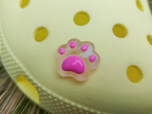 Glow-in-the-dark pastel paw print charms in pink, yellow, blue, and green, attached to a yellow shoe. The charms have a soft glow effect and add a playful animal-inspired look.