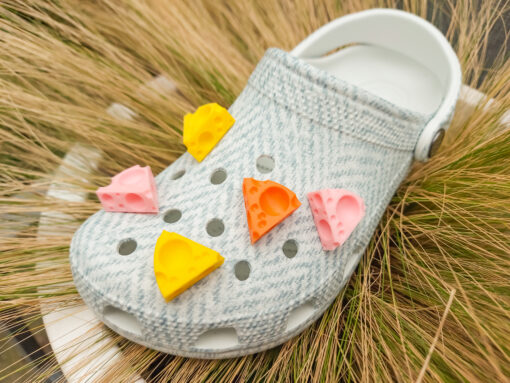 Close-up of a pair of light blue shoes decorated with colorful 3D-printed cheese wedge charms in shades of yellow, orange, and pink. The cheese charms feature realistic holes and a playful, quirky design, adding a fun pop of color to the footwear.