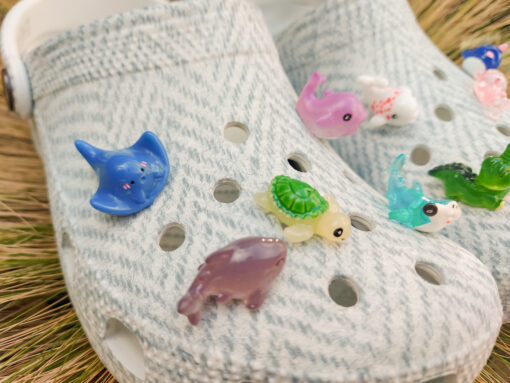 10-piece set of sea animal charms attached to a pair of woven-textured light gray shoes, displayed on a bed of dried grass. The collection includes colorful mini sea creatures like a turtle, shark, whale, narwhal, and starfish.