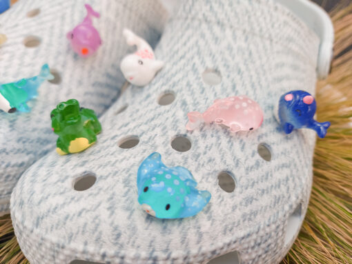 10-piece set of sea animal charms attached to a pair of woven-textured light gray shoes, displayed on a bed of dried grass. The collection includes colorful mini sea creatures like a turtle, shark, whale, narwhal, and starfish.