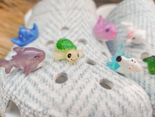 10-piece set of sea animal charms attached to a pair of woven-textured light gray shoes, displayed on a bed of dried grass. The collection includes colorful mini sea creatures like a turtle, shark, whale, narwhal, and starfish.