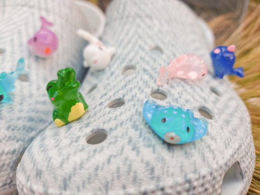 10-piece set of sea animal charms attached to a pair of woven-textured light gray shoes, displayed on a bed of dried grass. The collection includes colorful mini sea creatures like a turtle, shark, whale, narwhal, and starfish.