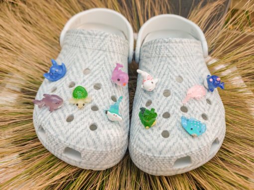 10-piece set of sea animal charms attached to a pair of woven-textured light gray shoes, displayed on a bed of dried grass. The collection includes colorful mini sea creatures like a turtle, shark, whale, narwhal, and starfish.