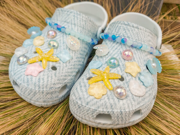 A 24-piece beach-themed charm and chain set on a pair of light gray woven-texture shoes. The set includes sea-inspired charms like starfish, shells, pearls, and iridescent accents with two beaded chains draped across the tops, evoking an oceanic look